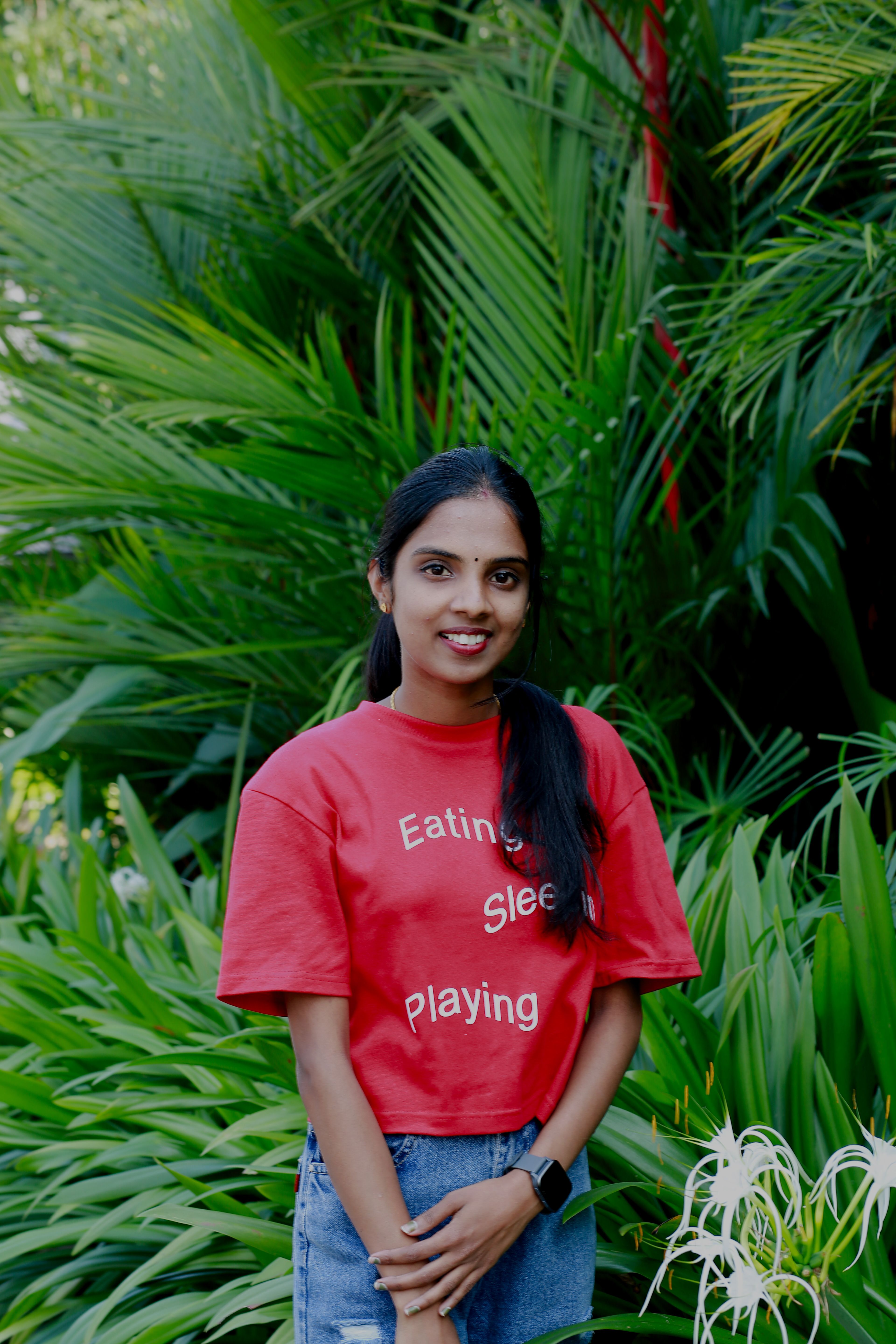 Smiling young woman in a red t-shirt stands amidst lush, vibrant greenery enjoying the outdoors.