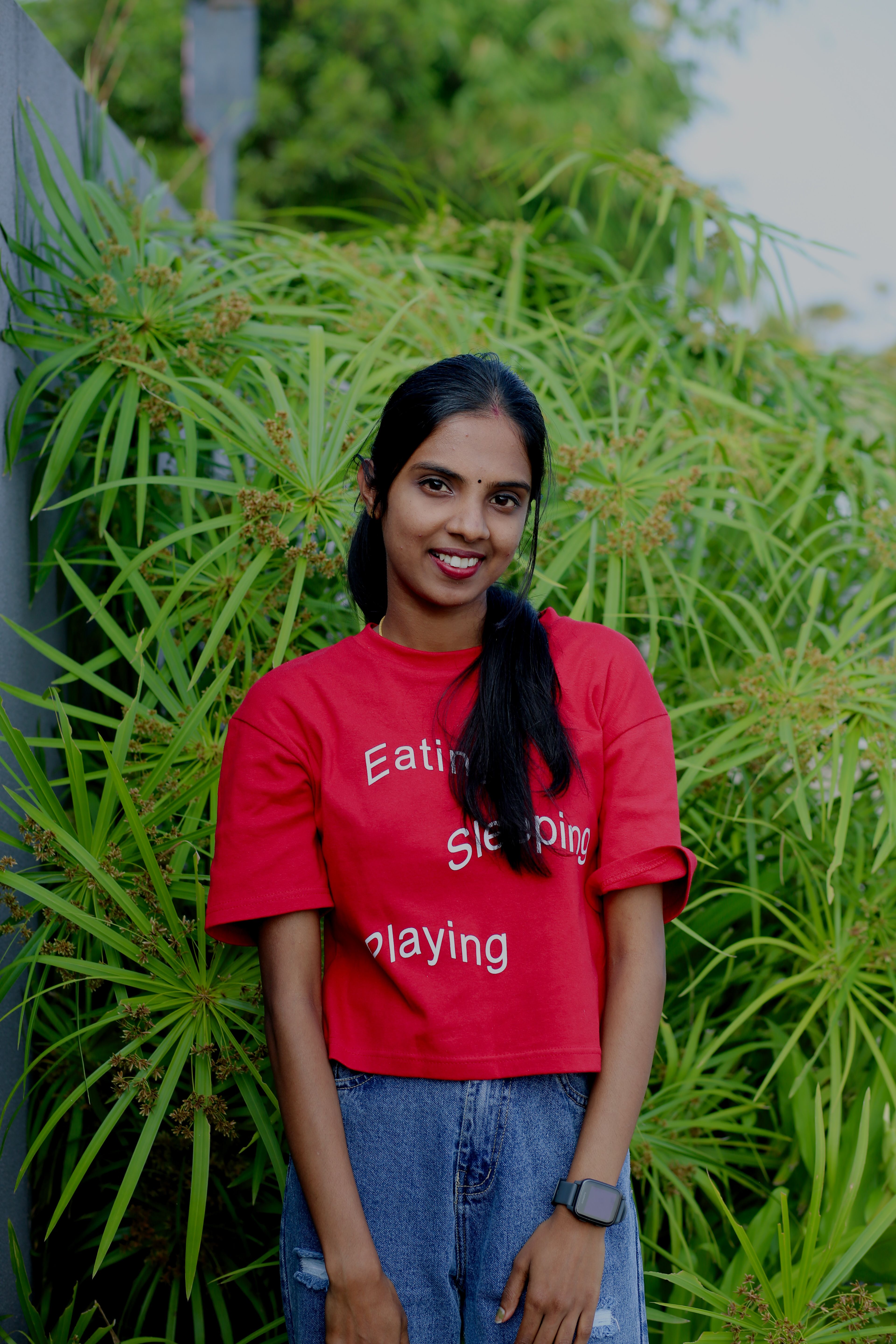 Smiling woman in a red t-shirt against a lush green plant background.