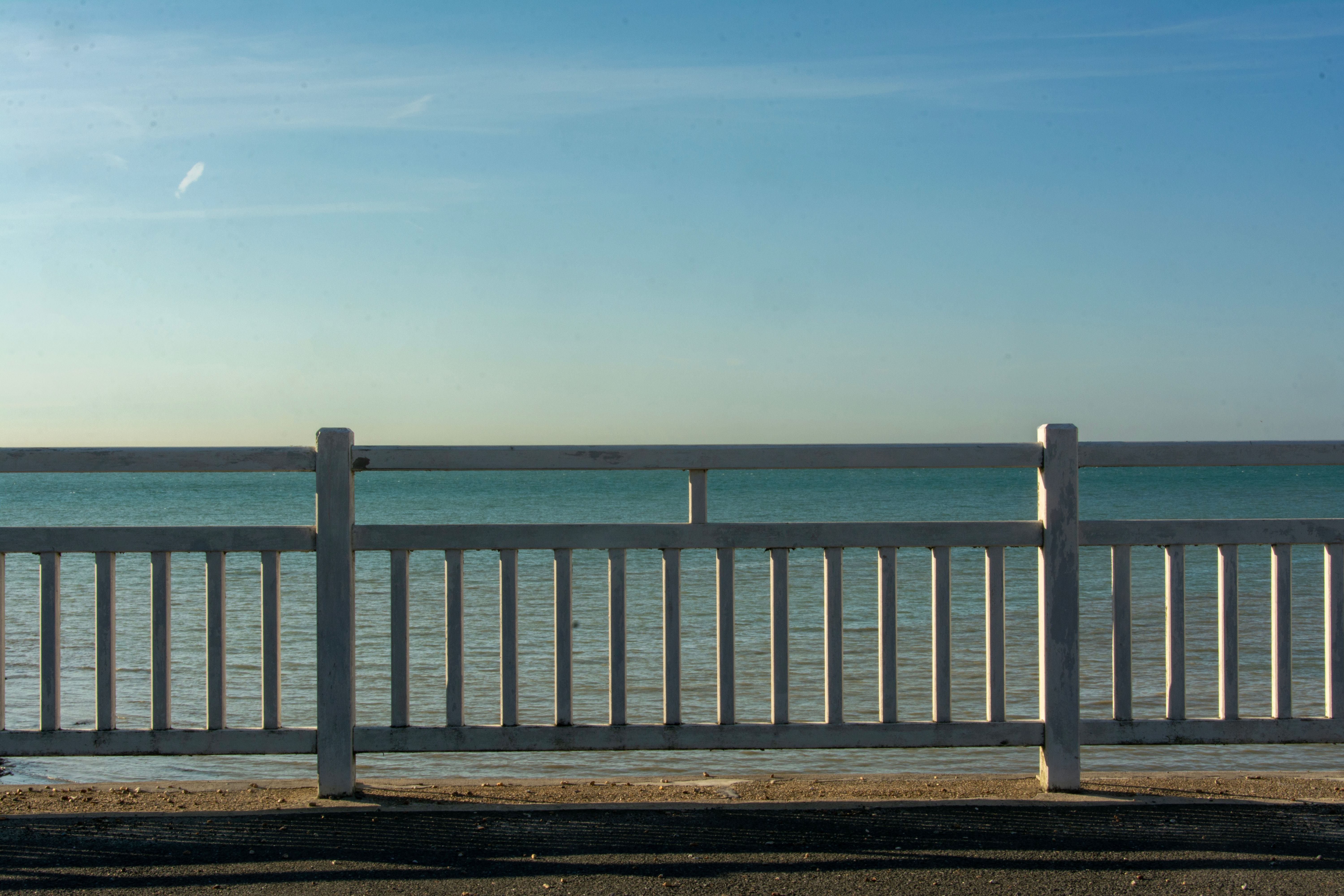 Peaceful seascape with a clear sky, viewed through a white wooden fence on a calm day.