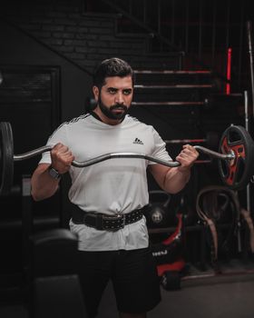Focused man performs barbell curl exercise in a dimly lit gym, showcasing fitness determination.
