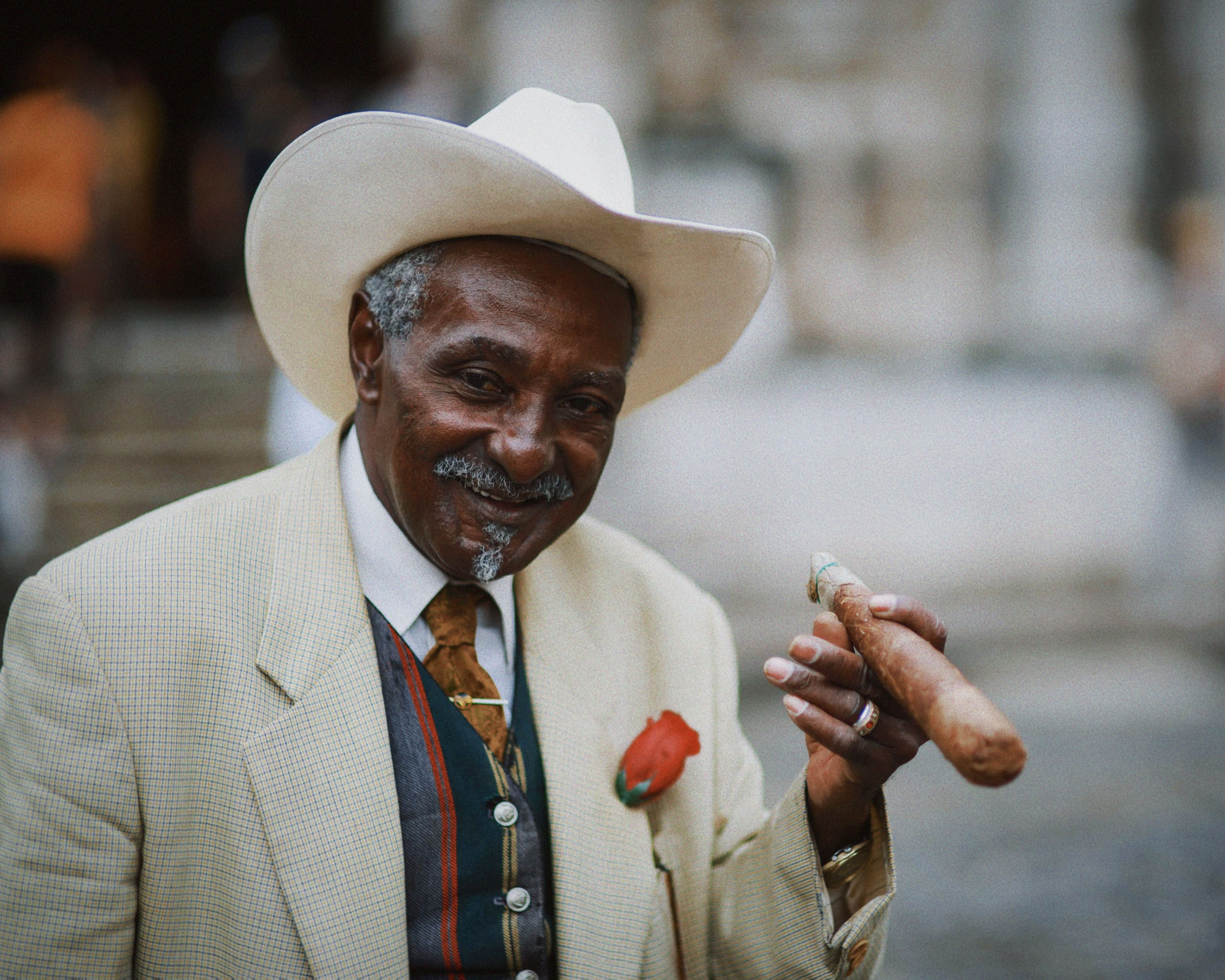 Elderly man in a suit and hat, smiling while holding a cigar outdoors. A warm and friendly portrait.