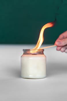 A hand lighting a white candle in a jar using a match, against a green background.