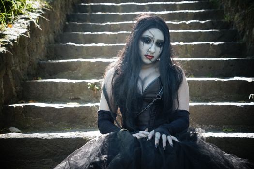 Mysterious woman in gothic attire sitting on ancient stone steps outdoors.