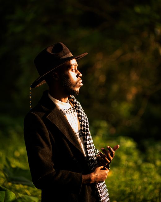 A fashionable man in a hat and scarf stands in a lush outdoor setting, illuminated by warm light.