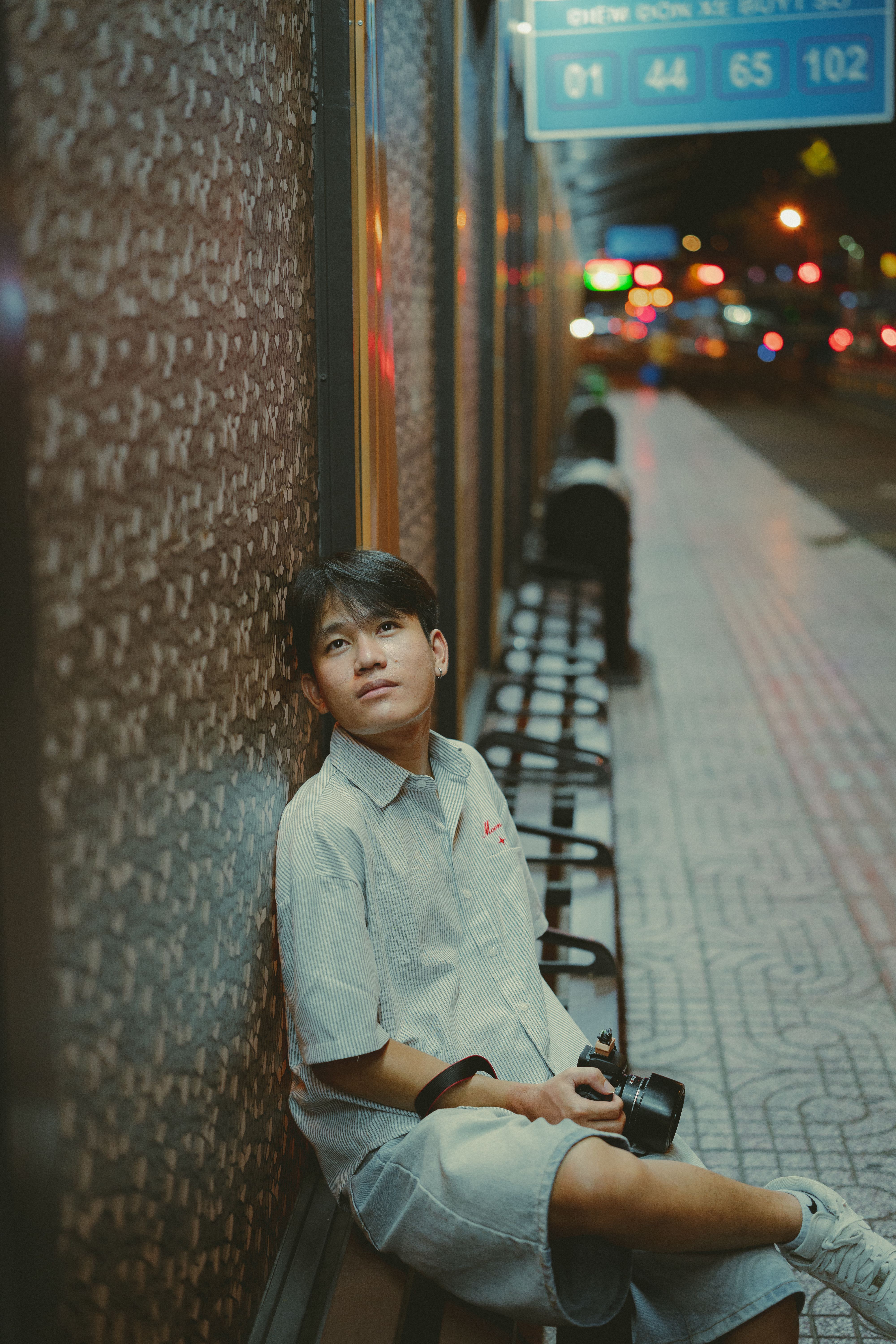 A young man sits leisurely on a city street in Ho Chi Minh City, Vietnam, with vibrant lights in the background.