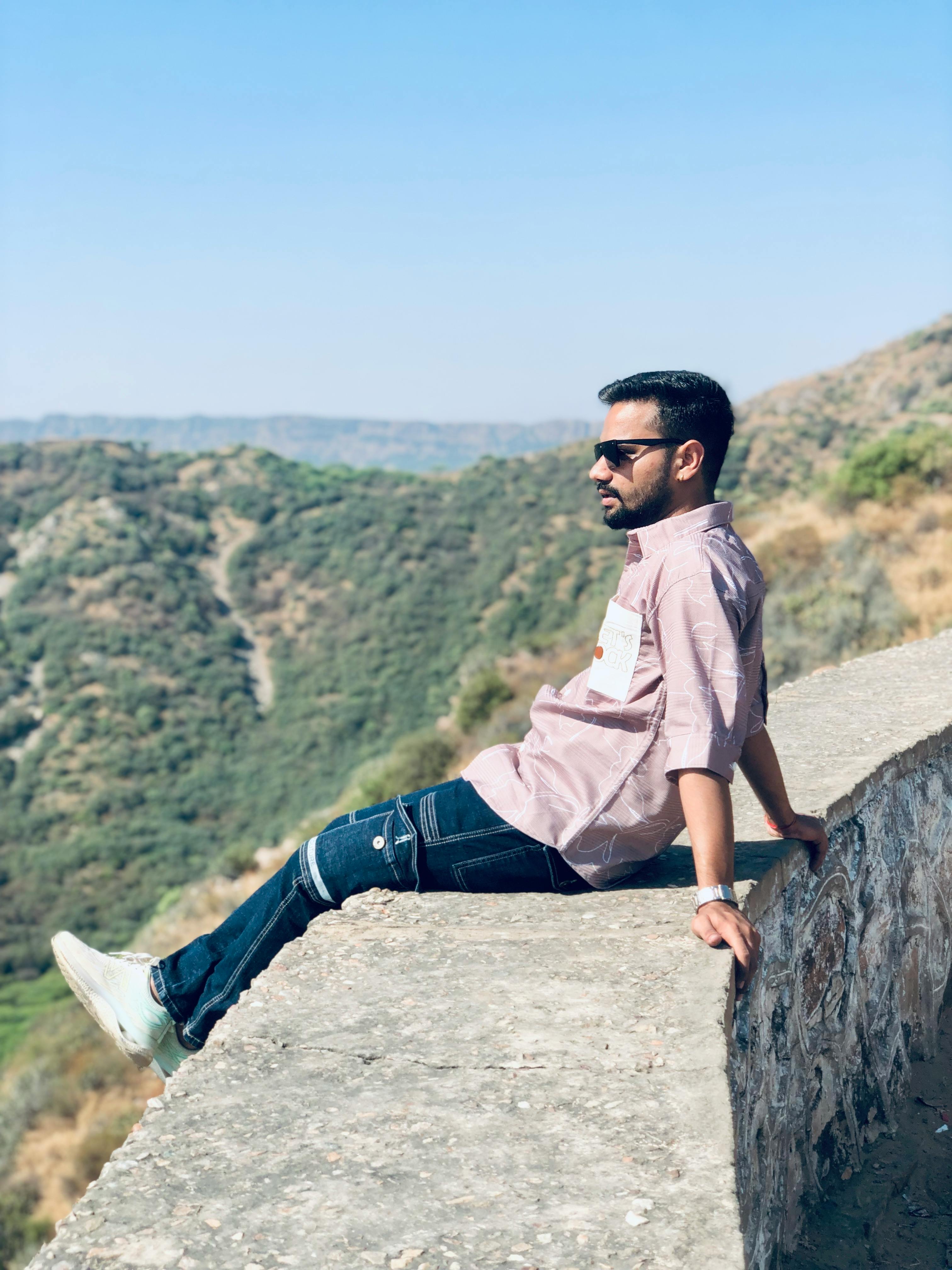 A man in casual attire sits on a stone wall, enjoying a scenic valley view on a sunny day.