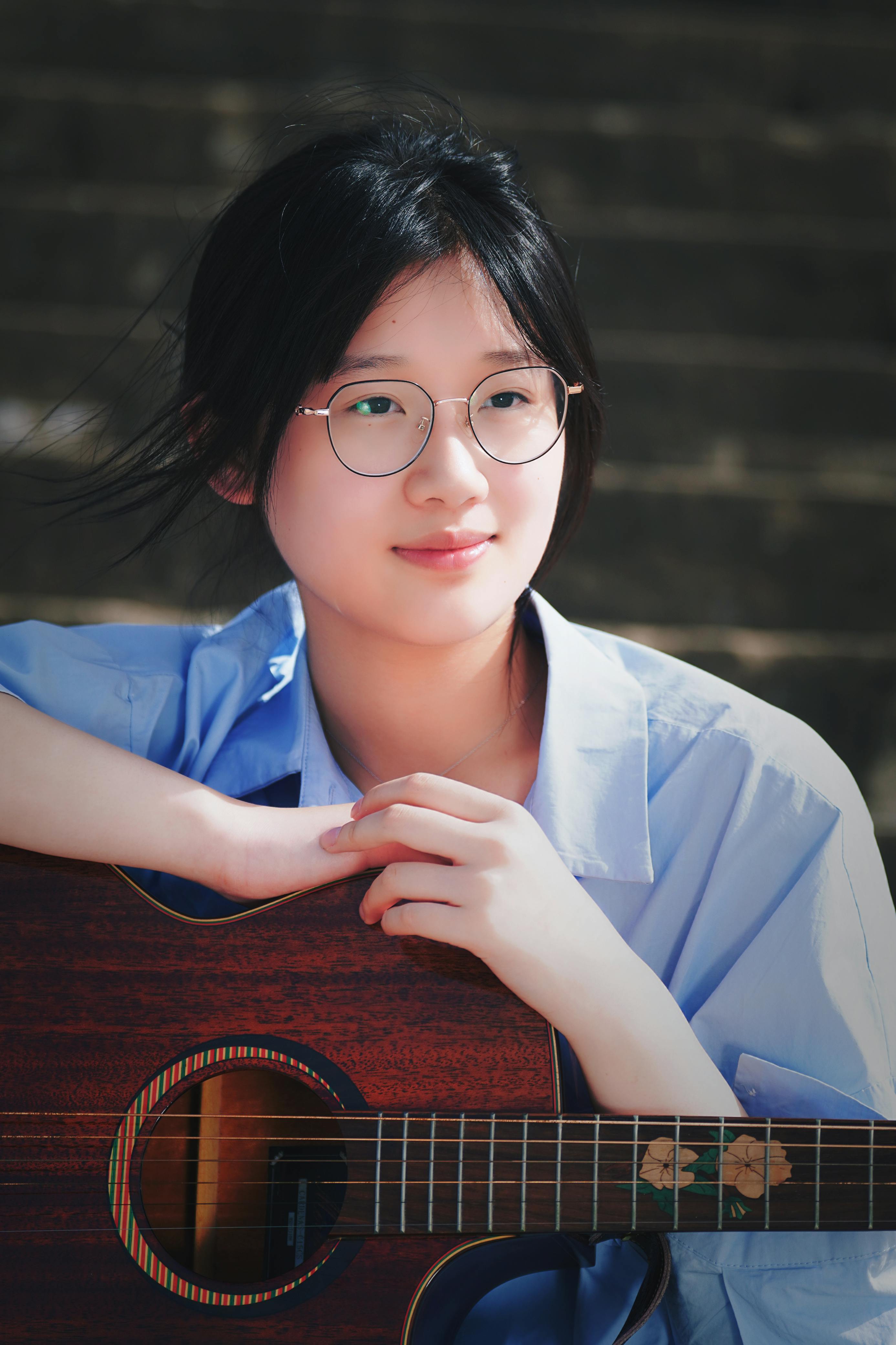 Young woman with glasses holding a guitar, enjoying a quiet moment outdoors. Ideal for music and lifestyle themes.