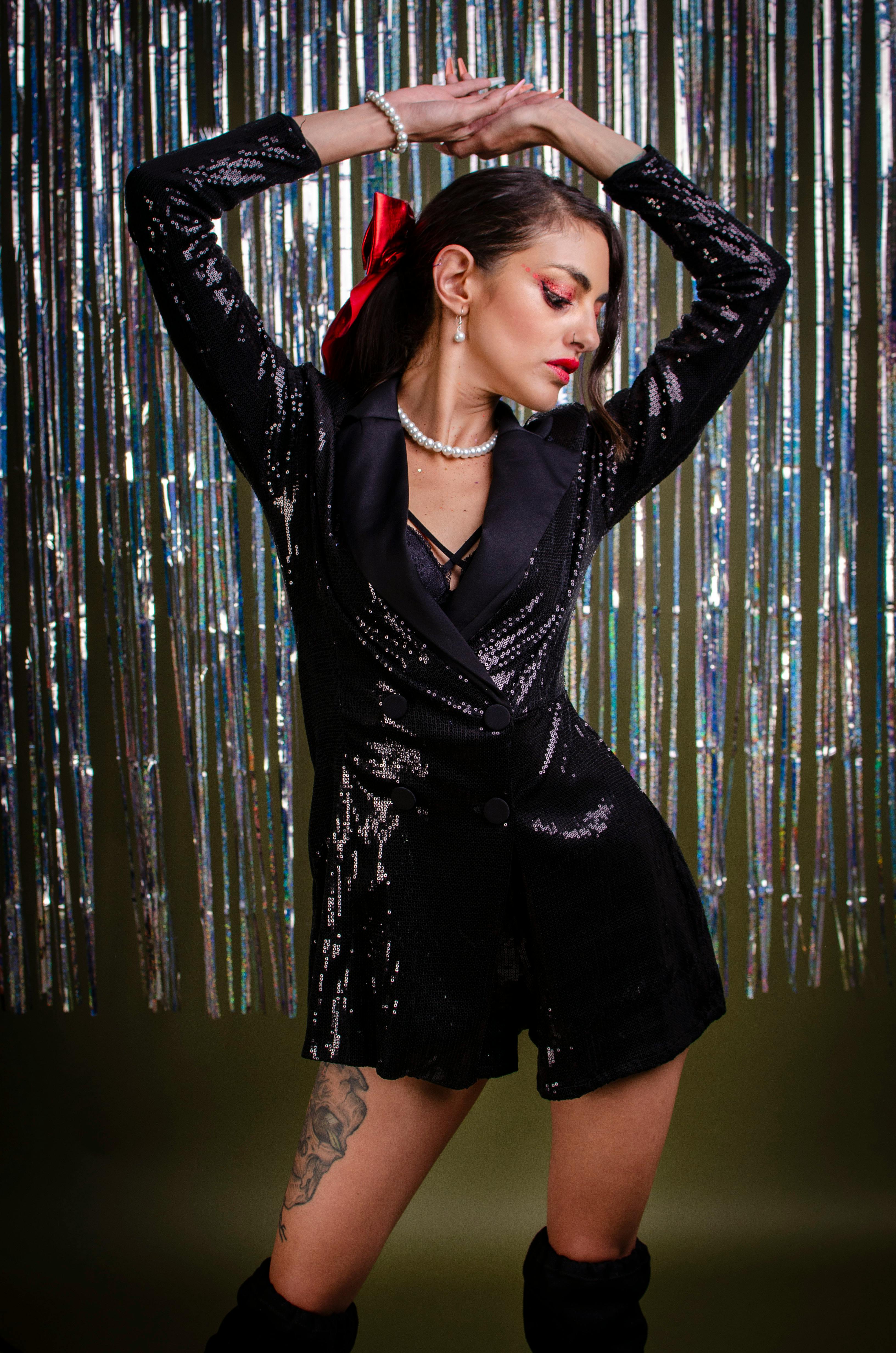 grátis Mulher estilosa em um vestido preto brilhante posando confiantemente em um ambiente interno com um fundo brilhante. Foto profissional