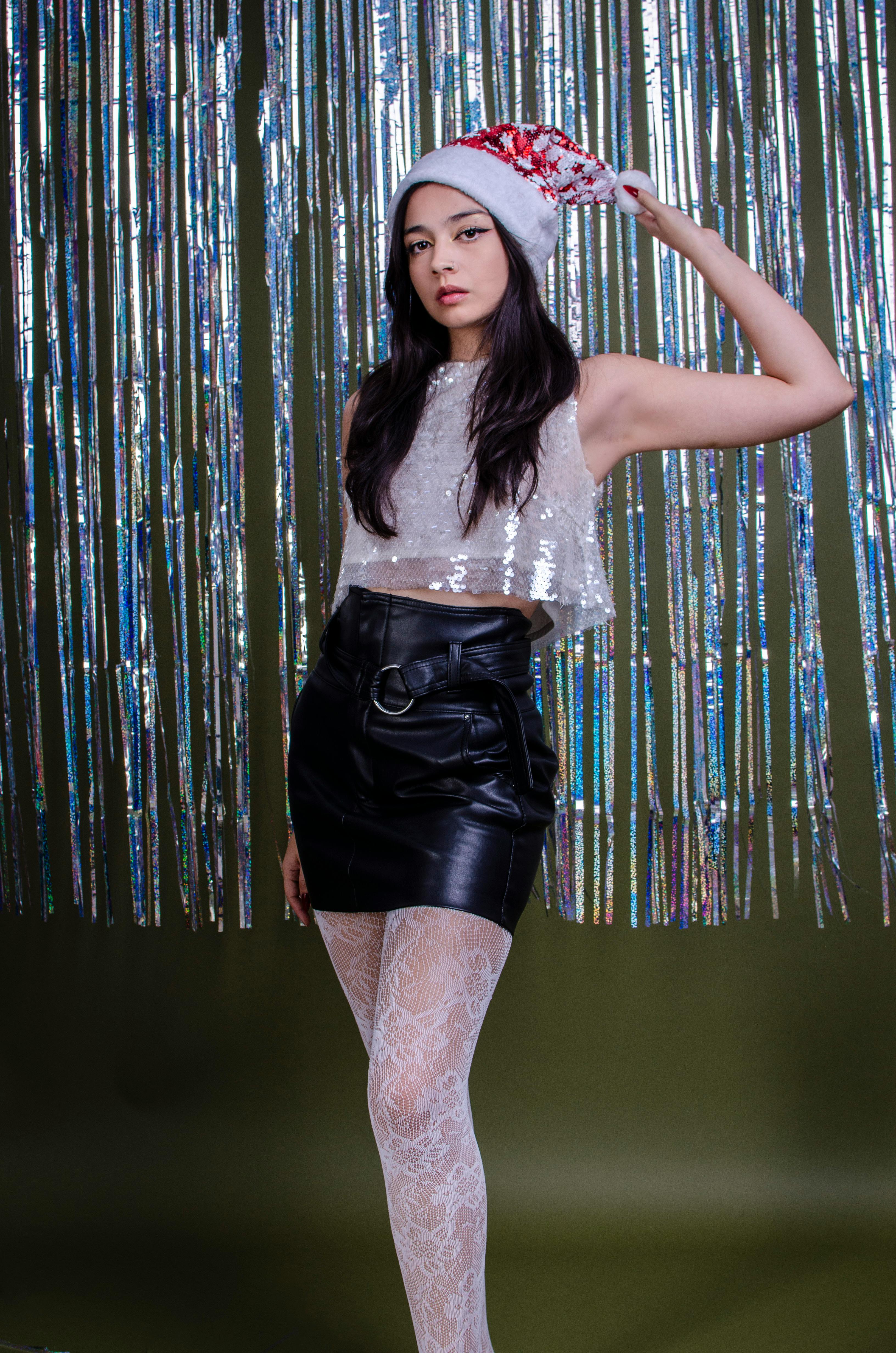 grátis Mulher estilosa com blusa de lantejoulas e chapéu de Papai Noel posando em um cenário festivo. Foto profissional