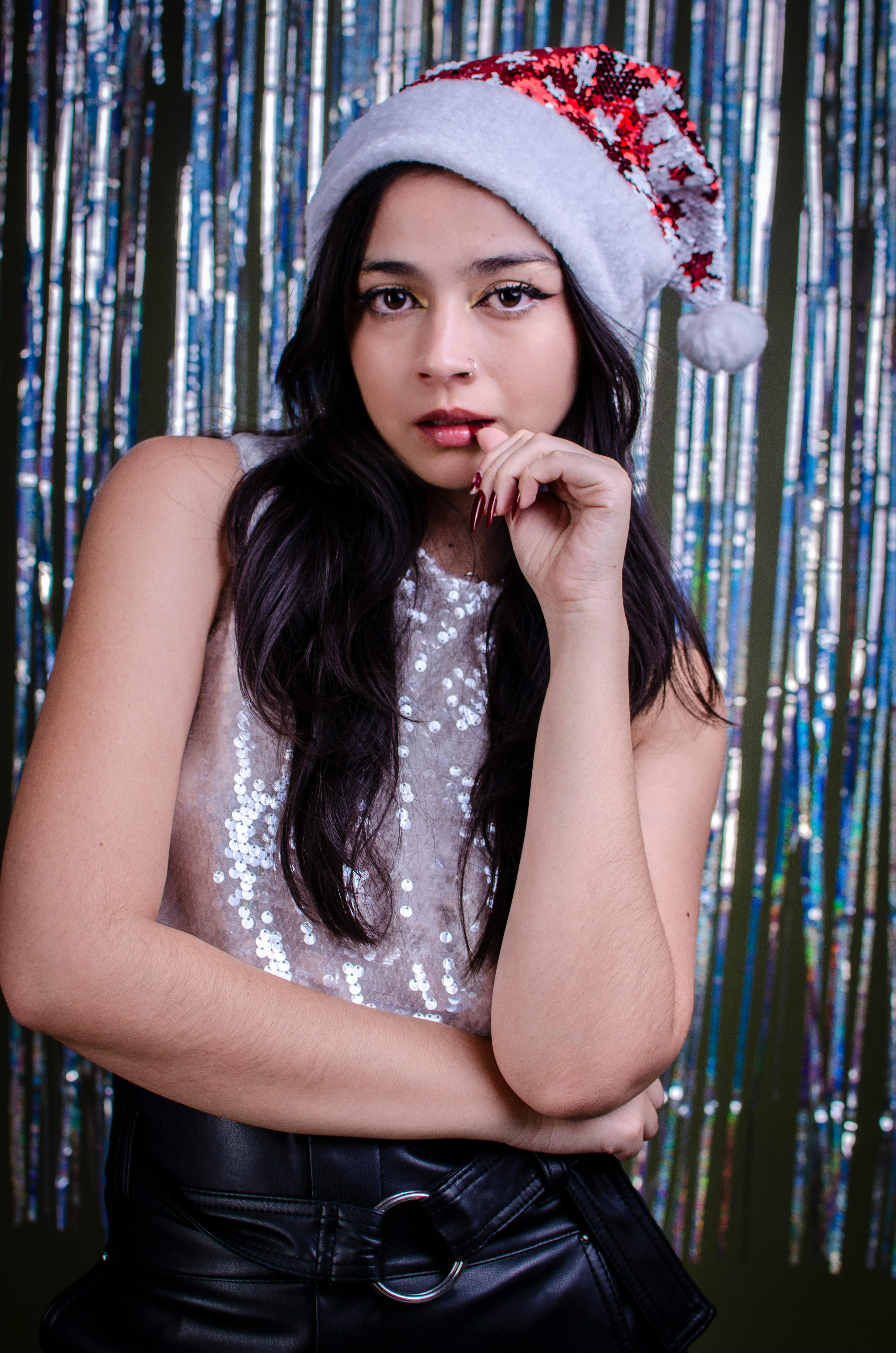 Gratis Una donna elegante posa in un interno con un cappello da Babbo Natale, aggiungendo un tocco natalizio al suo look elegante. Foto a disposizione