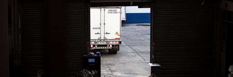 A white delivery truck exits a dimly lit warehouse loading dock, surrounded by packages and pallets.