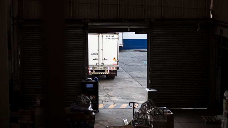 A white delivery truck exits a dimly lit warehouse loading dock, surrounded by packages and pallets.