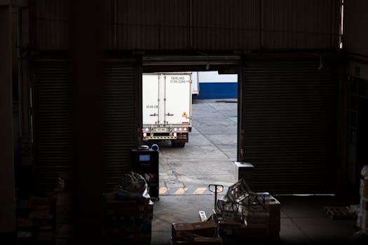 A white delivery truck exits a dimly lit warehouse loading dock, surrounded by packages and pallets.