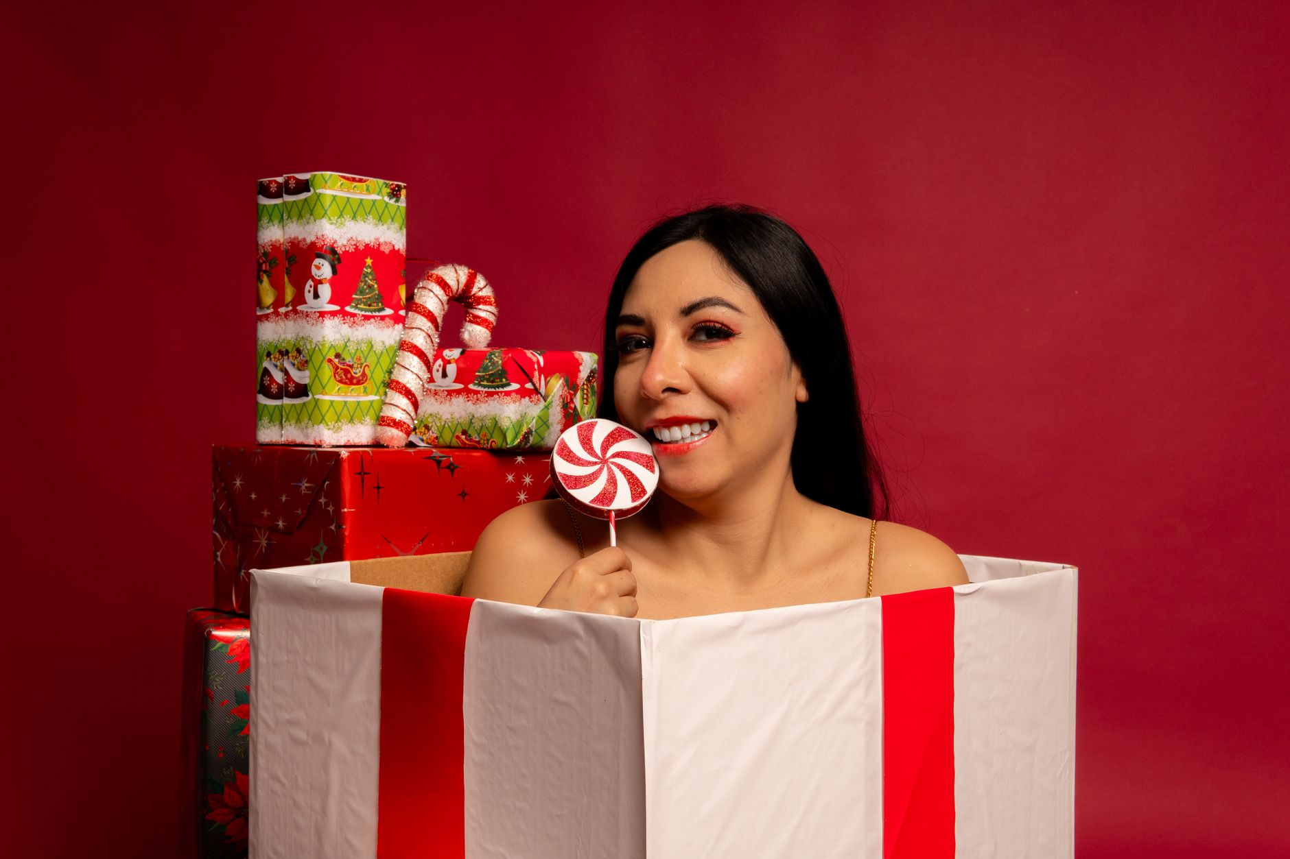 A joyful Mexican woman posing in a gift box with holiday candy against a red background.