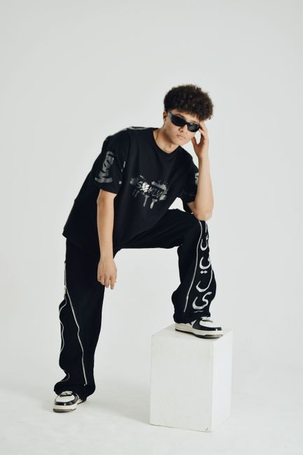 A young male model posing in trendy black attire against a white background in a studio setting.
