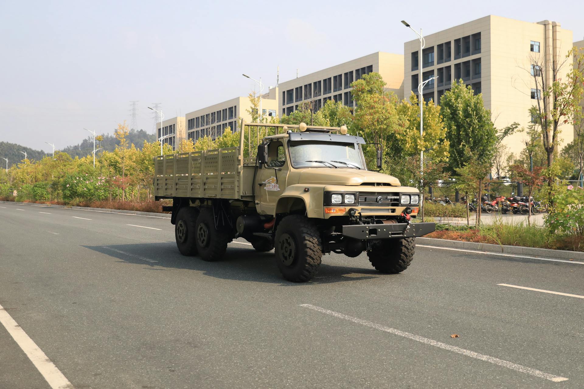 Tan military truck driving on a city road with autumn foliage and buildings in the background.