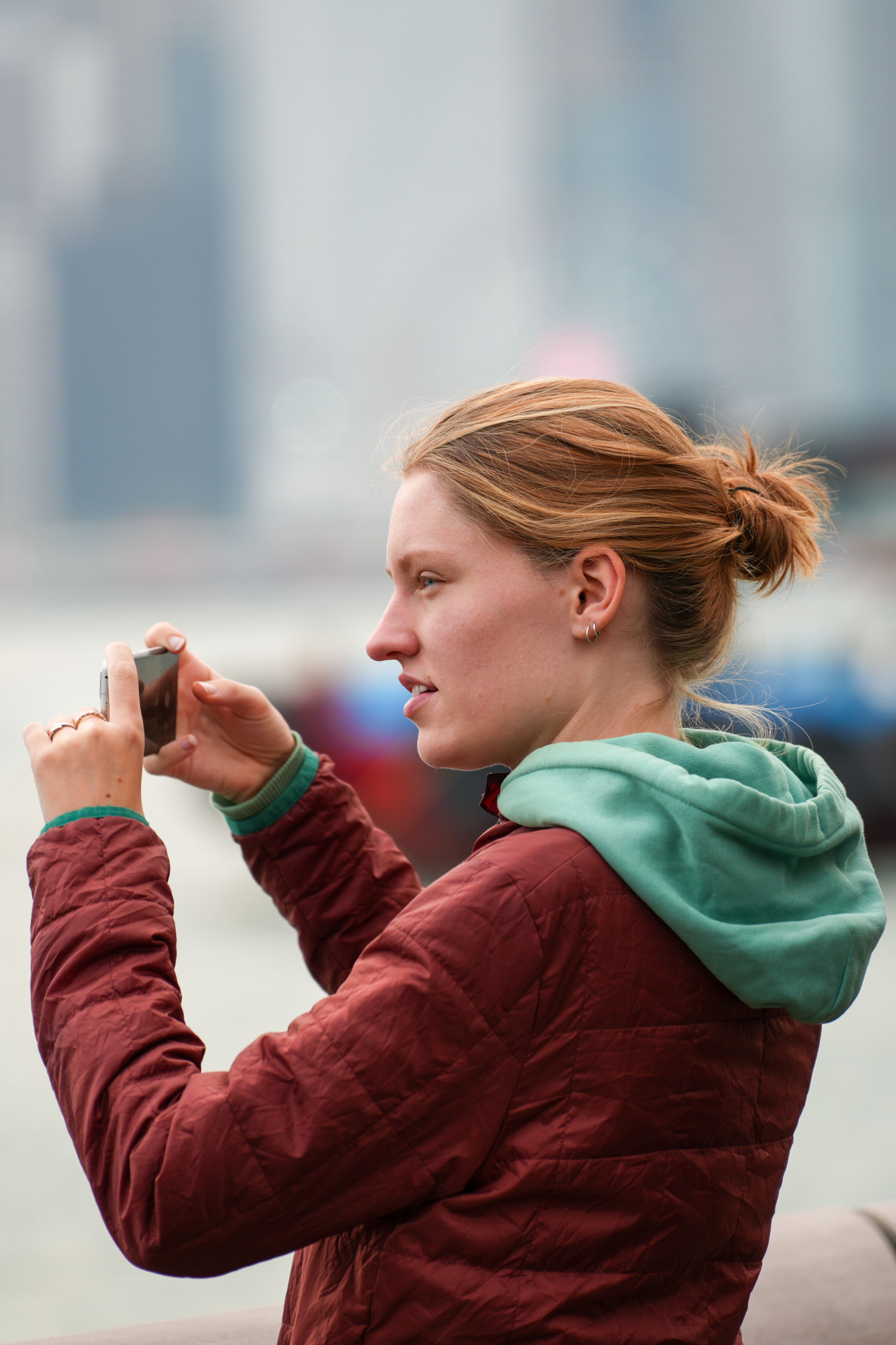 A woman taking a photo with her smartphone by the waterfront, city skyline blurred in the background.