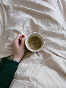 A cozy morning setting with a coffee cup held over beige sheets, evoking relaxation.