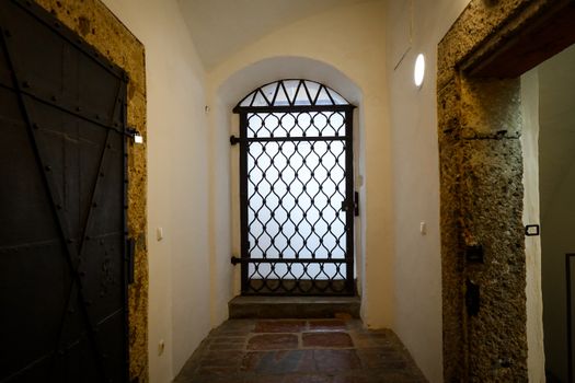 A dimly lit stone corridor featuring a fortified steel window with intricate design.