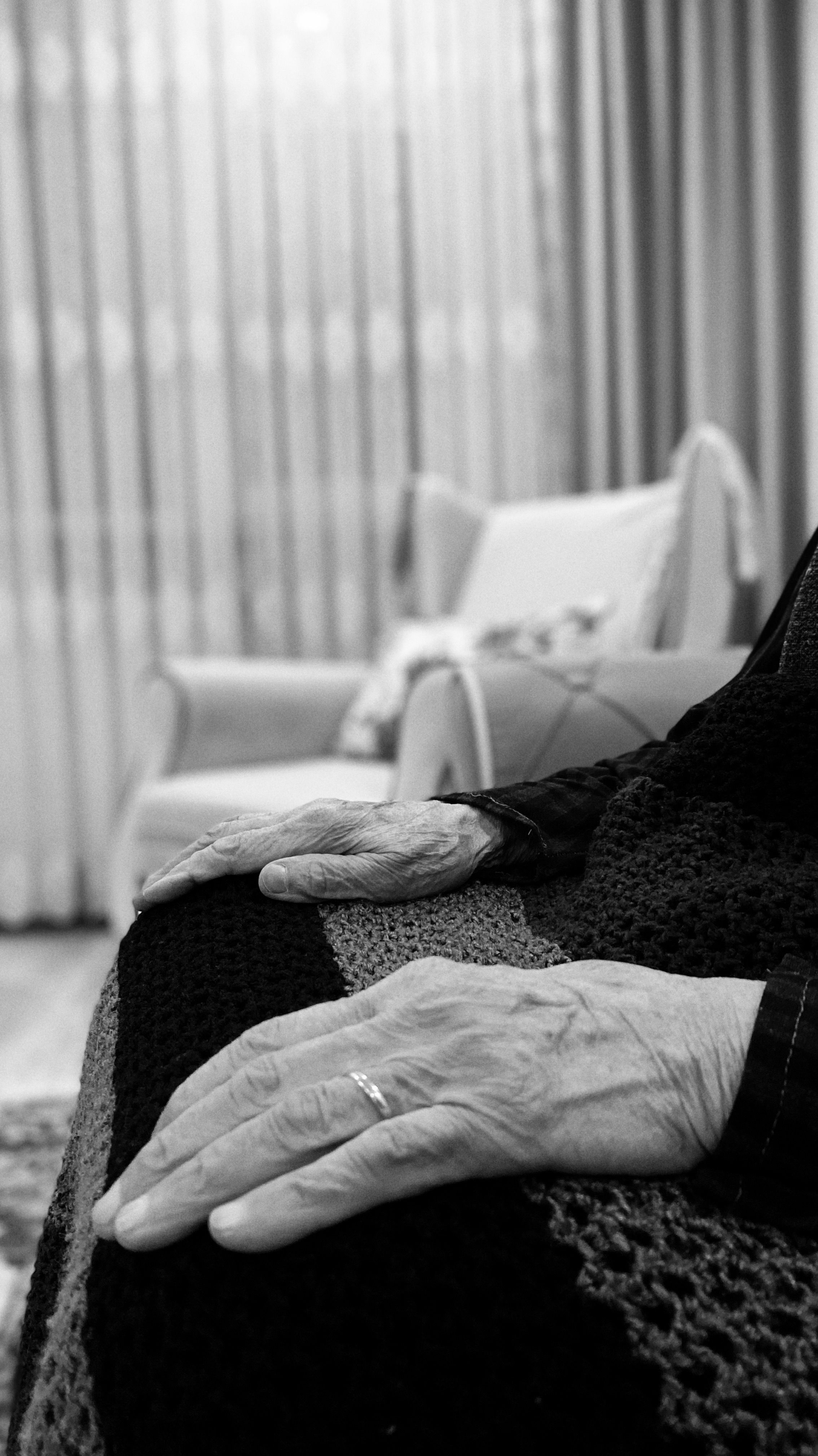 Black and white image of elderly hands resting on a crocheted blanket in a cozy living room.