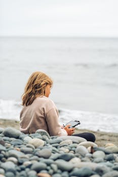 A woman operates a drone controller while seated on a pebble beach in British Columbia.