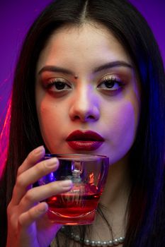 Captivating portrait of a woman with bold makeup holding a glass, lit by vibrant colors.
