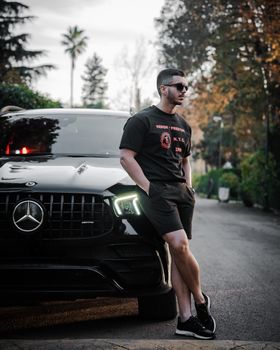 Young man in sunglasses leaning against a stylish black SUV on a street with trees.