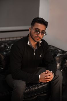Portrait of a young man in glasses sitting on a leather sofa with a serious expression.