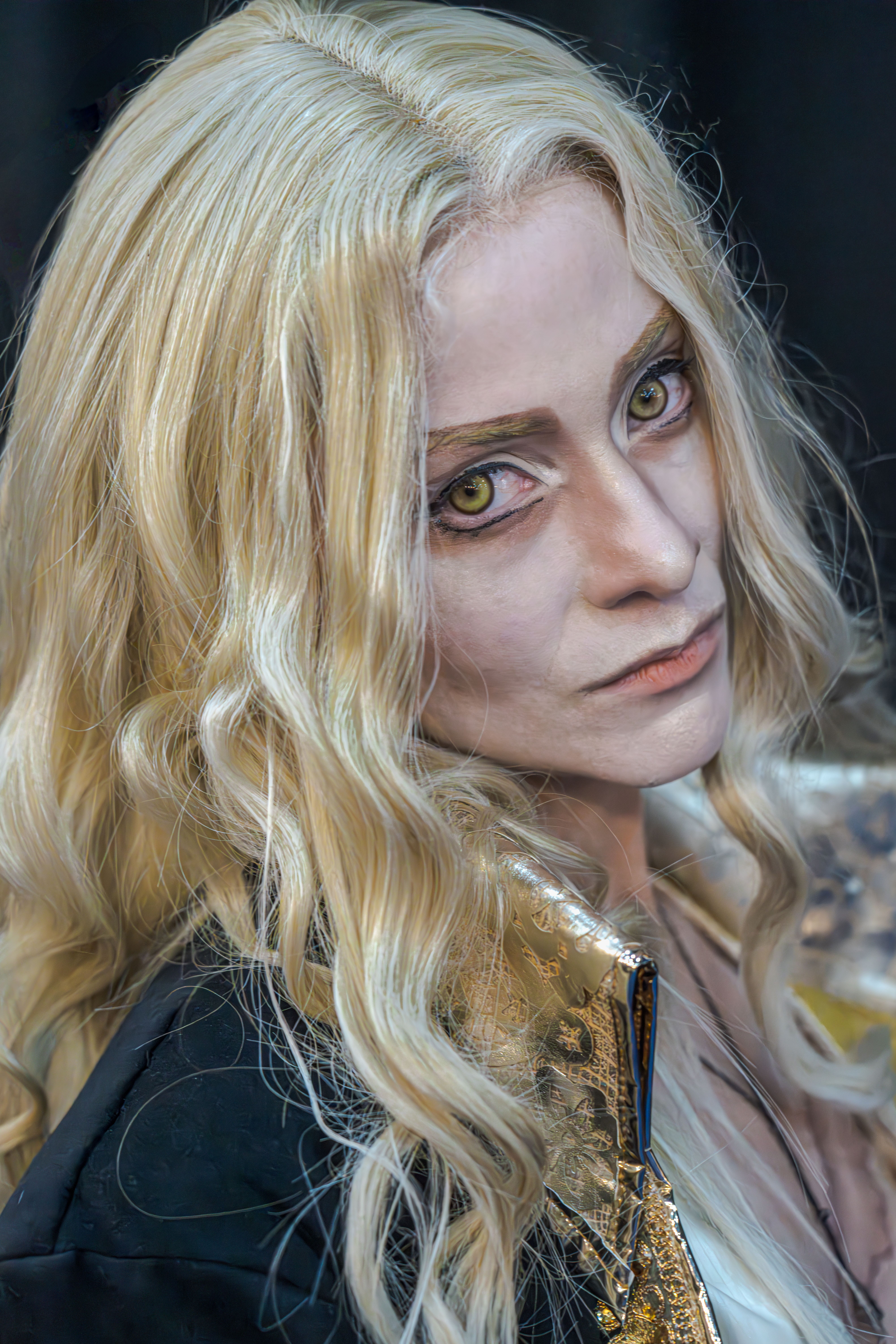Close-up portrait of a cosplayer with gothic and vampire makeup, intense gold eyes, and long blond hair.