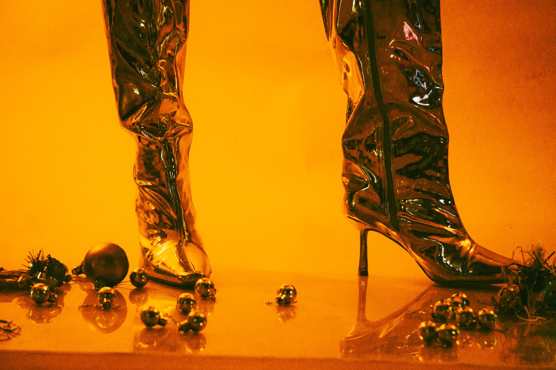 Shiny metallic high-heeled boots surrounded by festive holiday ornaments, set in a warm yellow ambiance.