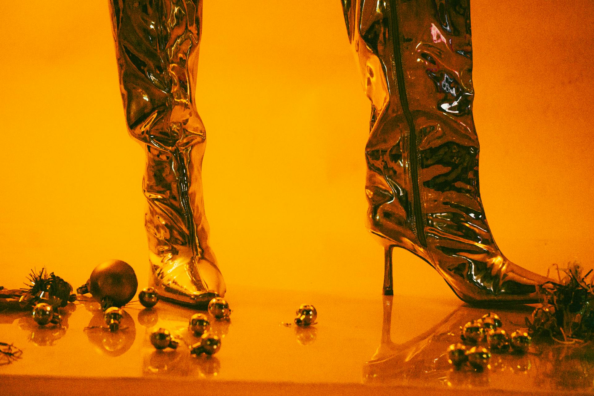 Shiny metallic high-heeled boots surrounded by festive holiday ornaments, set in a warm yellow ambiance.