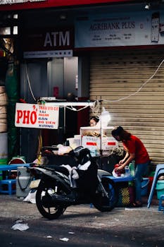 Street food vendor selling pho next to a motorbike on a busy street, creating a lively urban scene.