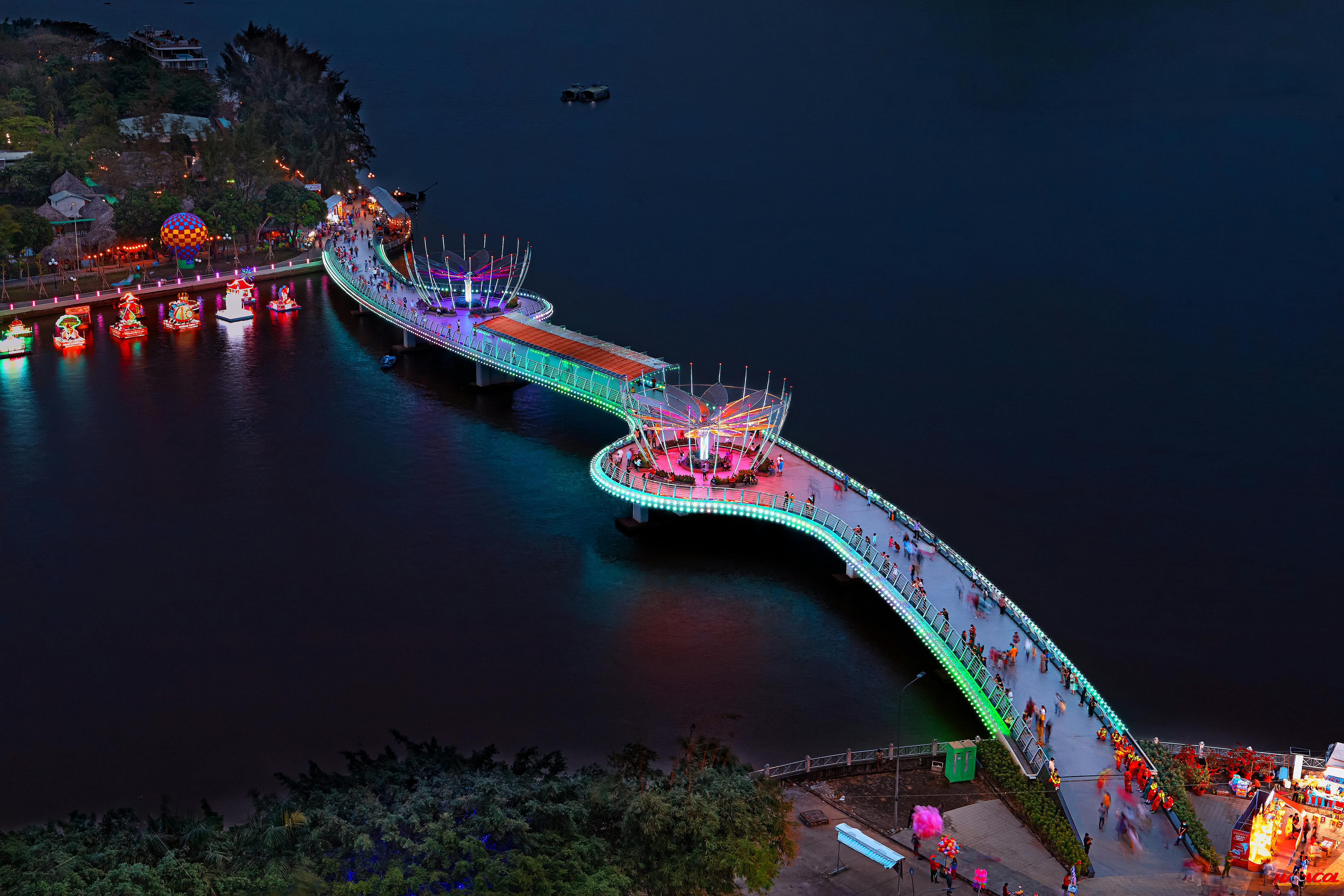 Aerial view of the illuminated pedestrian bridge over the river in Cu1ea7n Thu01a1, Vietnam at night, showcasing colorful lights.