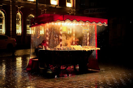 Rain-soaked street food vendor booth illuminated at night, offering grilled corn and chestnuts.