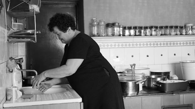 Black and white image of a woman washing dishes in a vintage kitchen setting, evoking nostalgia.