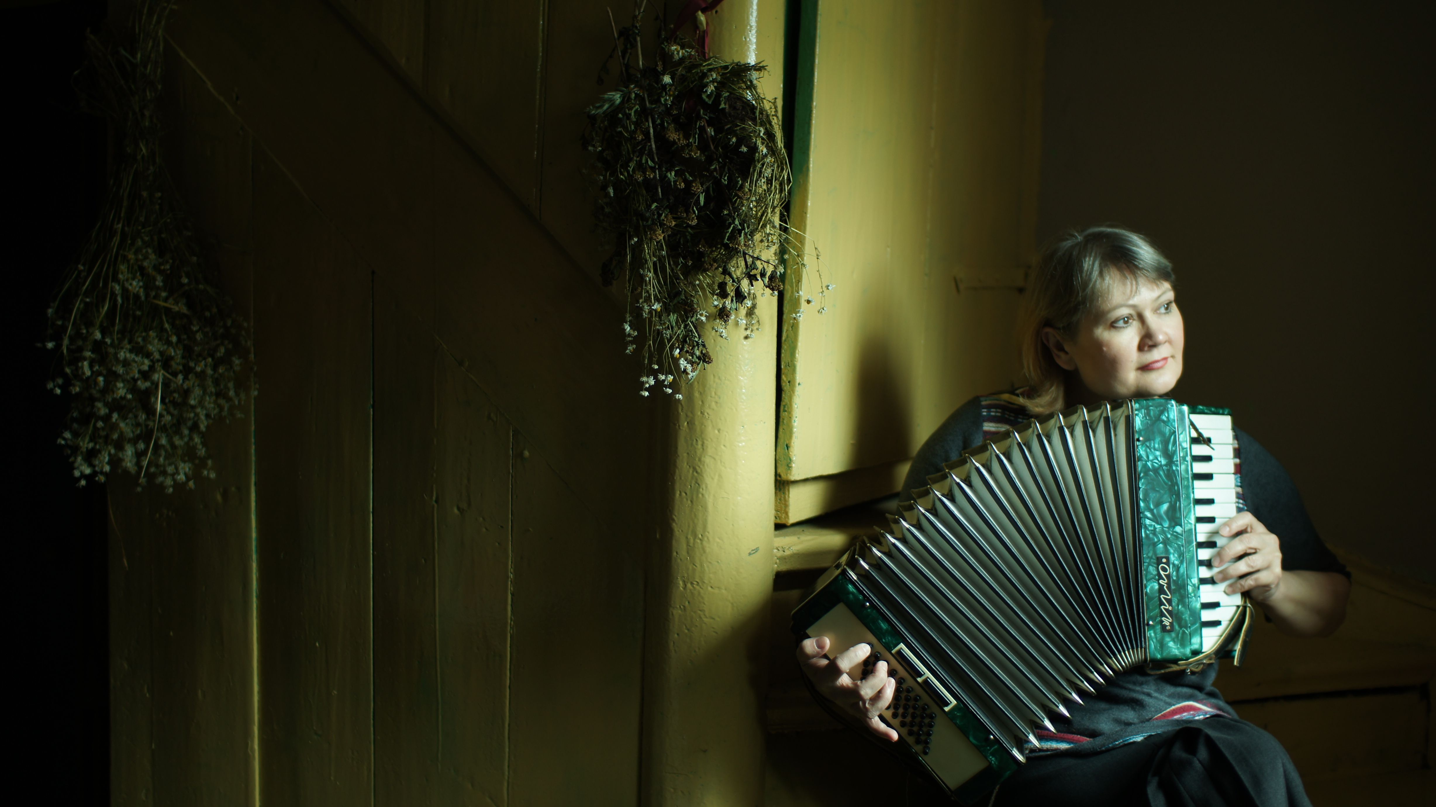 A woman playing an accordion indoors next to a wooden wall with dried flowers.