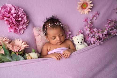 Free A peaceful baby sleeps among pink flowers and a stuffed bear, wrapped in soft lavender fabric. Stock Photo