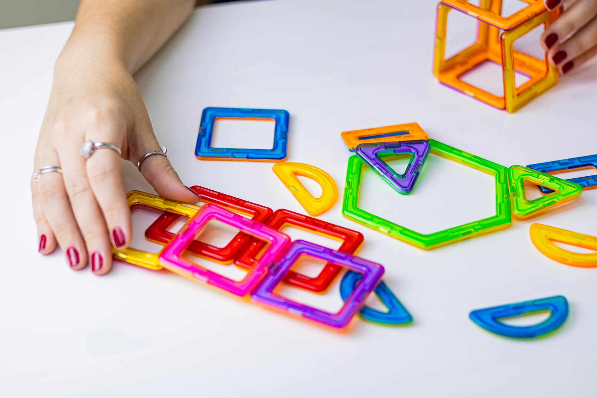 Hands playing with colorful geometric magnetic building blocks on a white surface.