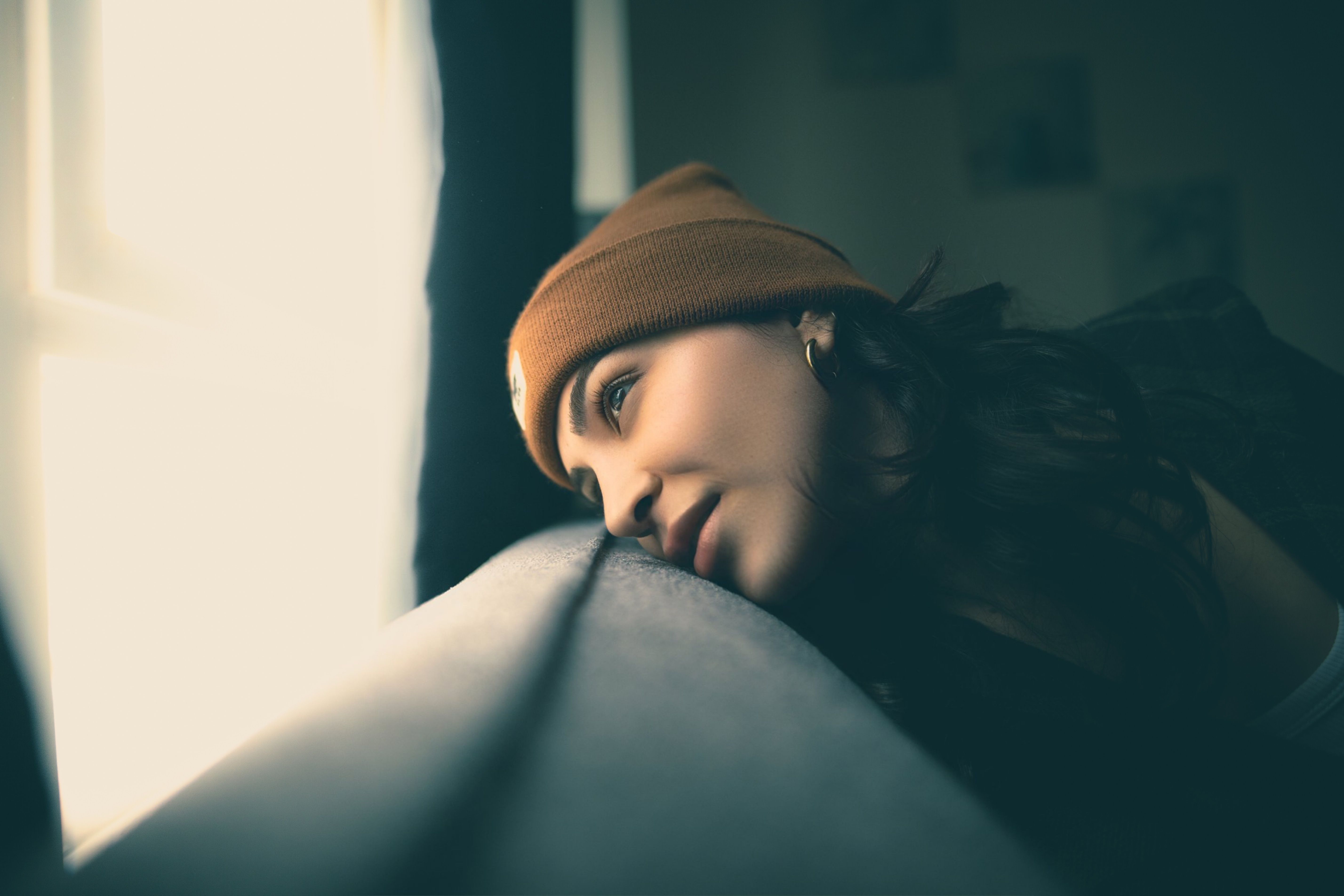 A thoughtful woman wearing a beanie gazes out of a sunlit window, immersed in deep contemplation.