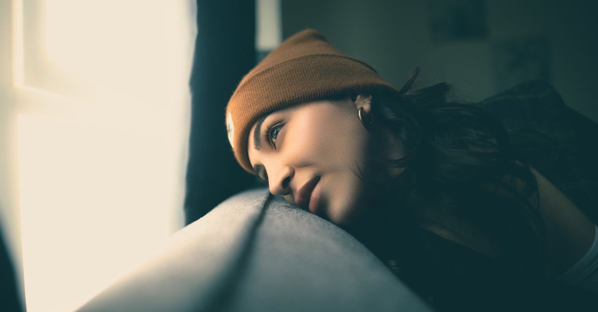 A thoughtful woman wearing a beanie gazes out of a sunlit window, immersed in deep contemplation.
