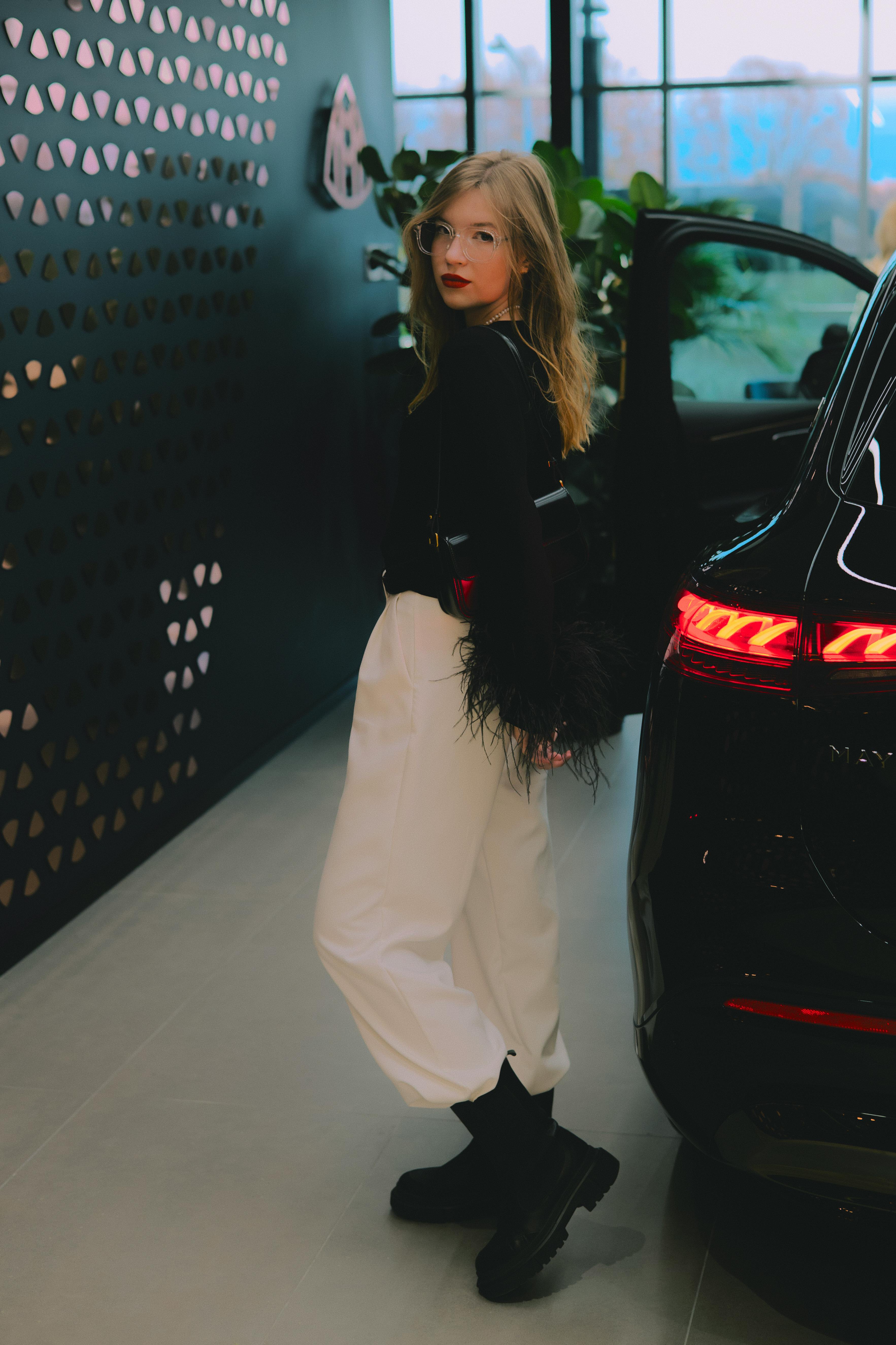 Gratis Wanita bergaya dengan pakaian monokrom berdiri di dekat mobil mewah di sebuah dealer, merangkul mode modern. Foto Stok