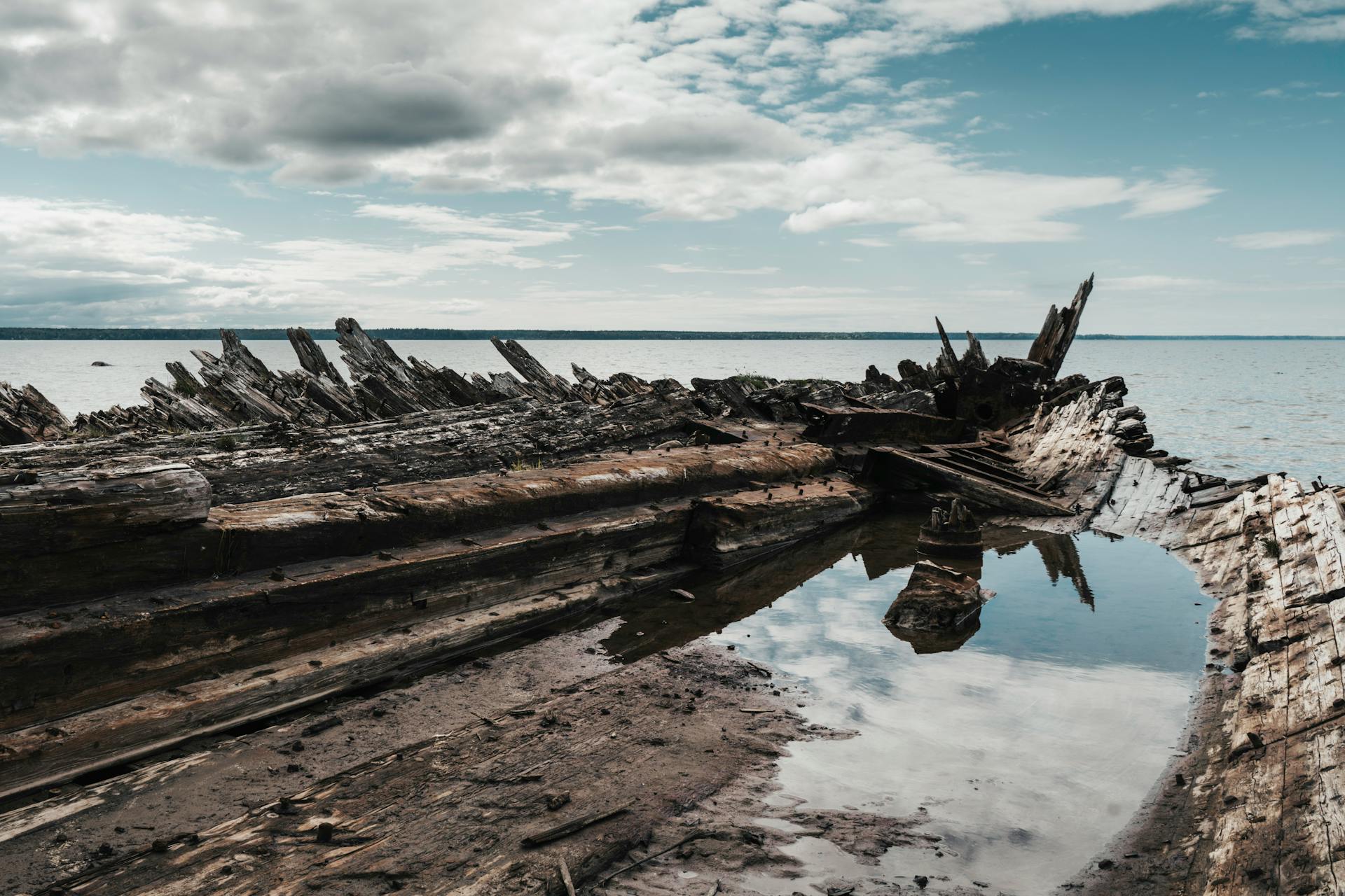 Aged wooden shipwreck on a sandy beach under a cloudy sky, reflecting in water.