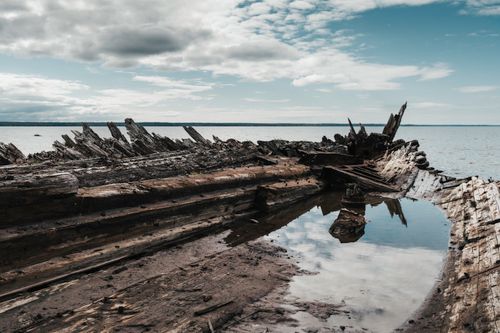 Free Aged wooden shipwreck on a sandy beach under a cloudy sky, reflecting in water. Stock Photo