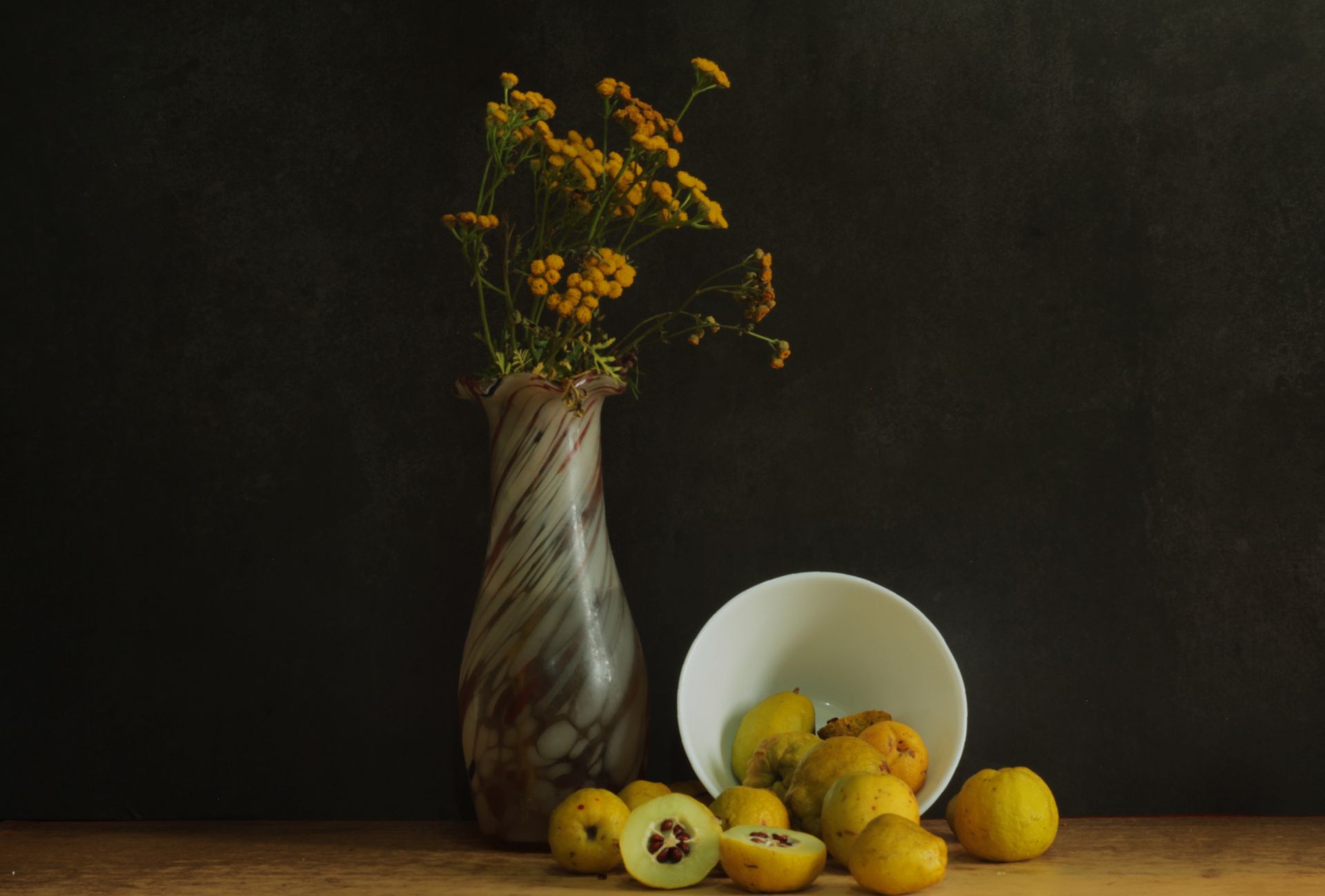 A still life photo featuring a vase with yellow flowers and quince fruits on a dark background.