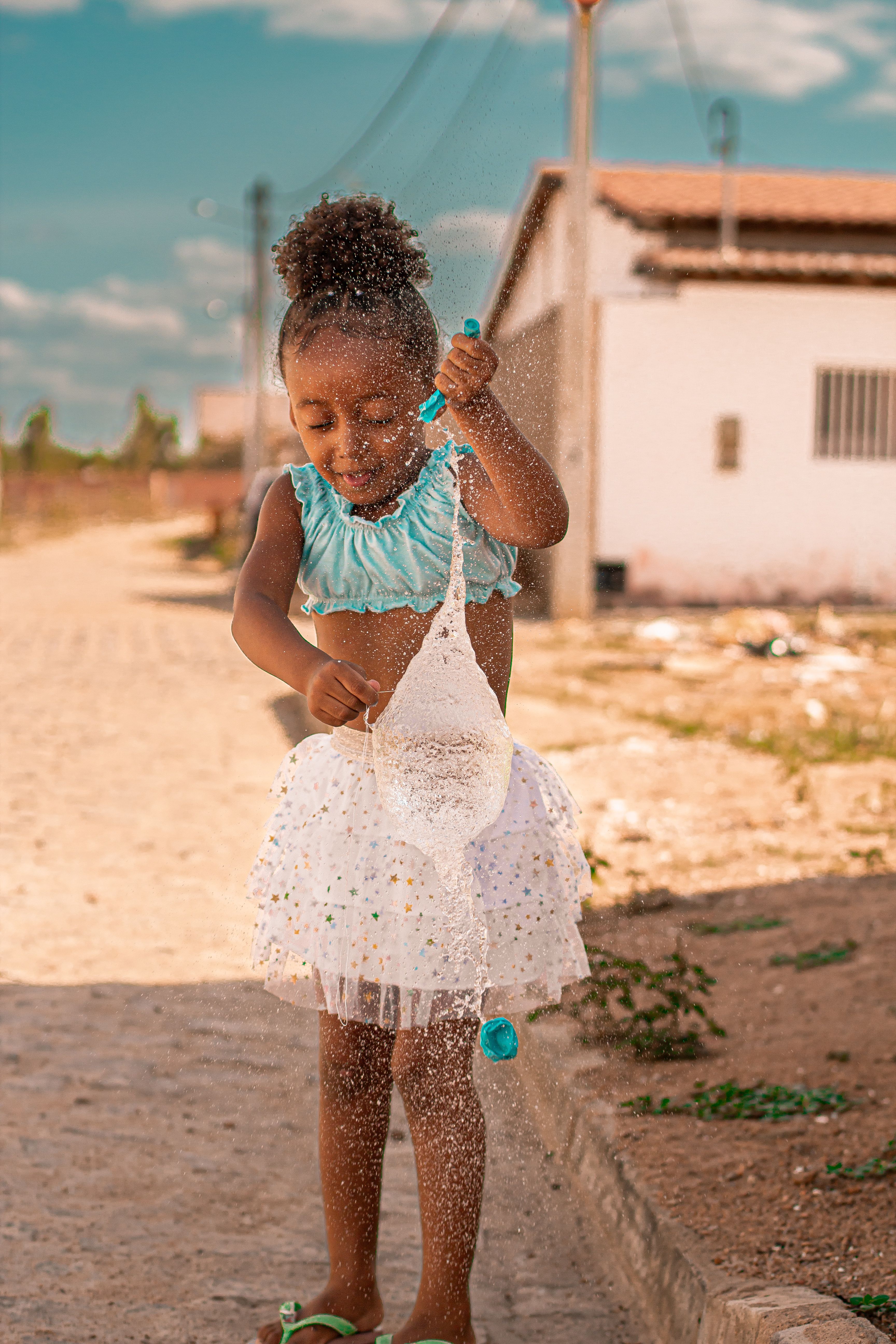 A happy child wearing a summer dress joyfully plays with water balloons on a sunny day.