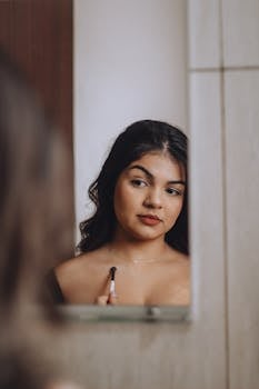 A woman is reflected in a mirror while applying makeup with a thoughtful expression.