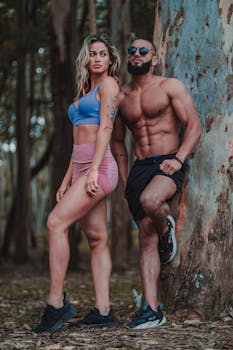 A fit couple posing confidently by a tree in a forest setting.