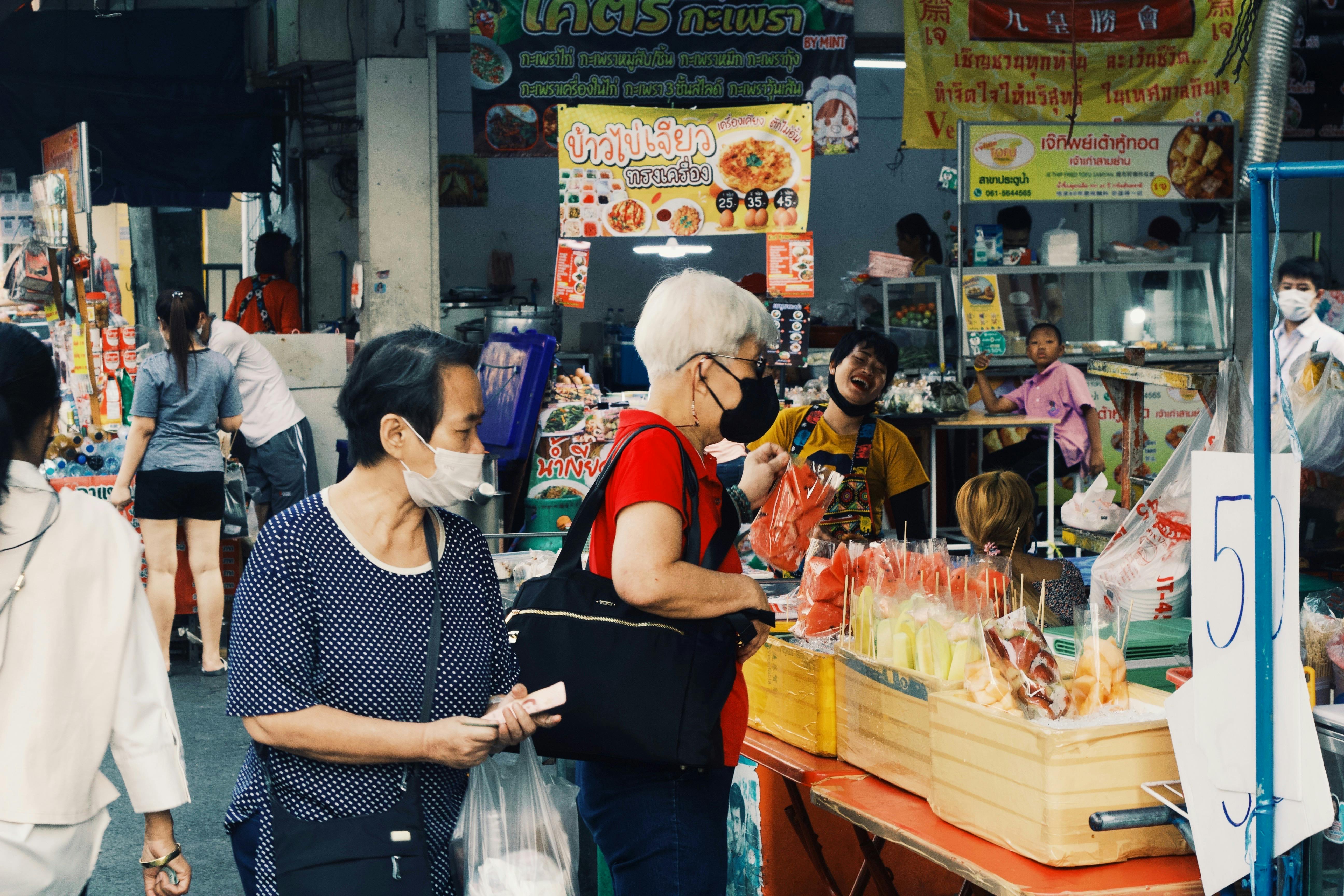 Free Colorful street market with vendors and shoppers wearing masks in bustling outdoor setting. Stock Photo