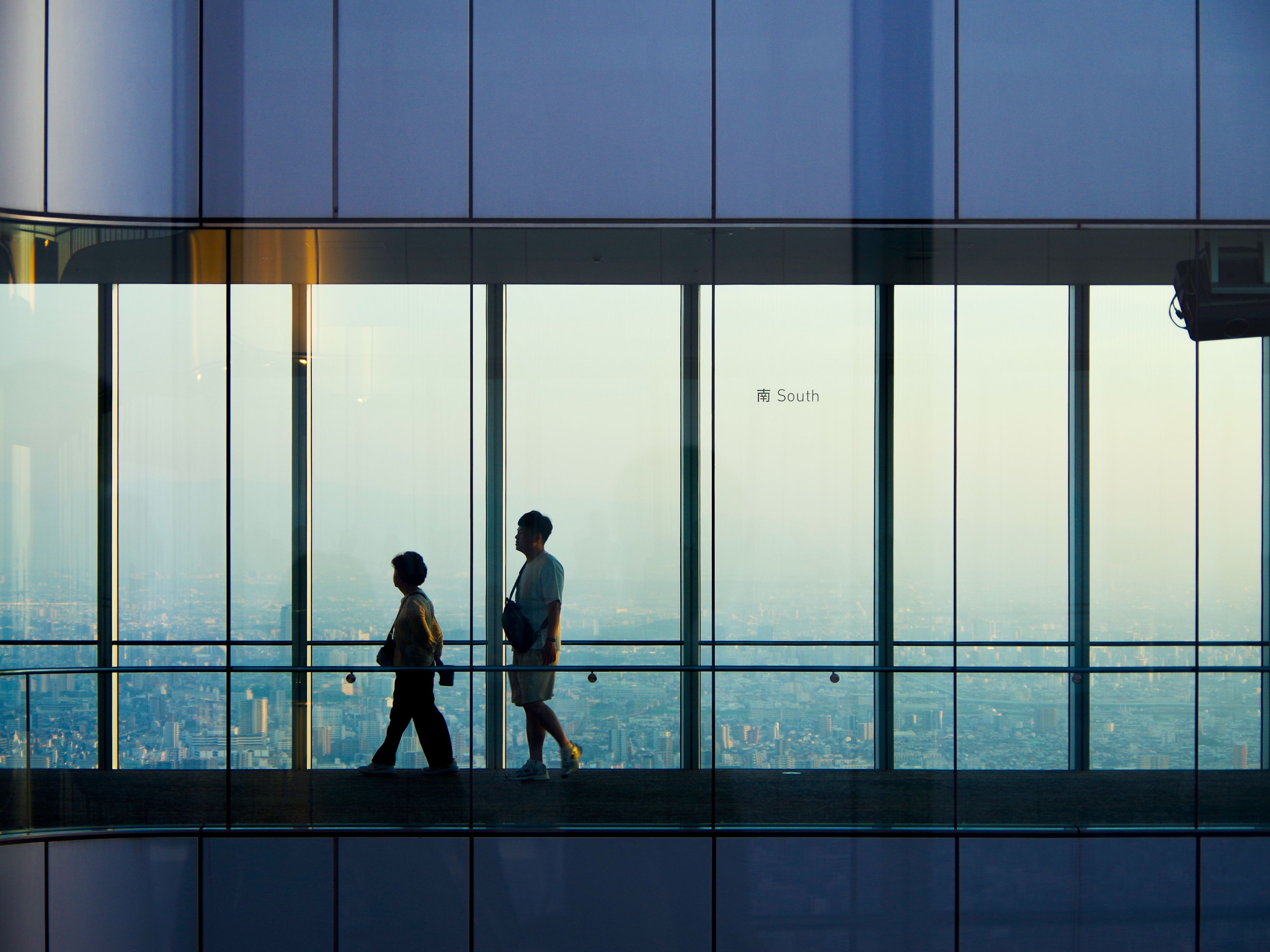 都市の景色を眺めながらガラスの廊下を歩く二人のシルエット。