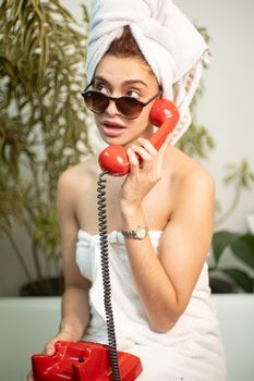 Woman with a towel on her head using a red vintage phone indoors, exuding a retro vibe.